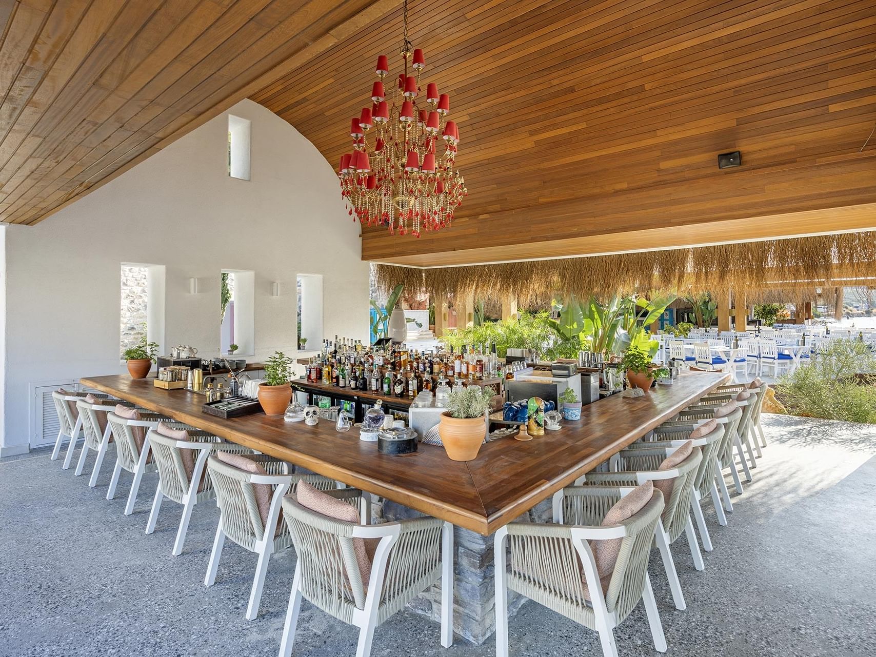 An outdoor restaurant at Titanic Luxury Collection Bodrum, featuring a wooden ceiling bar, a long table, and a red chandelier.