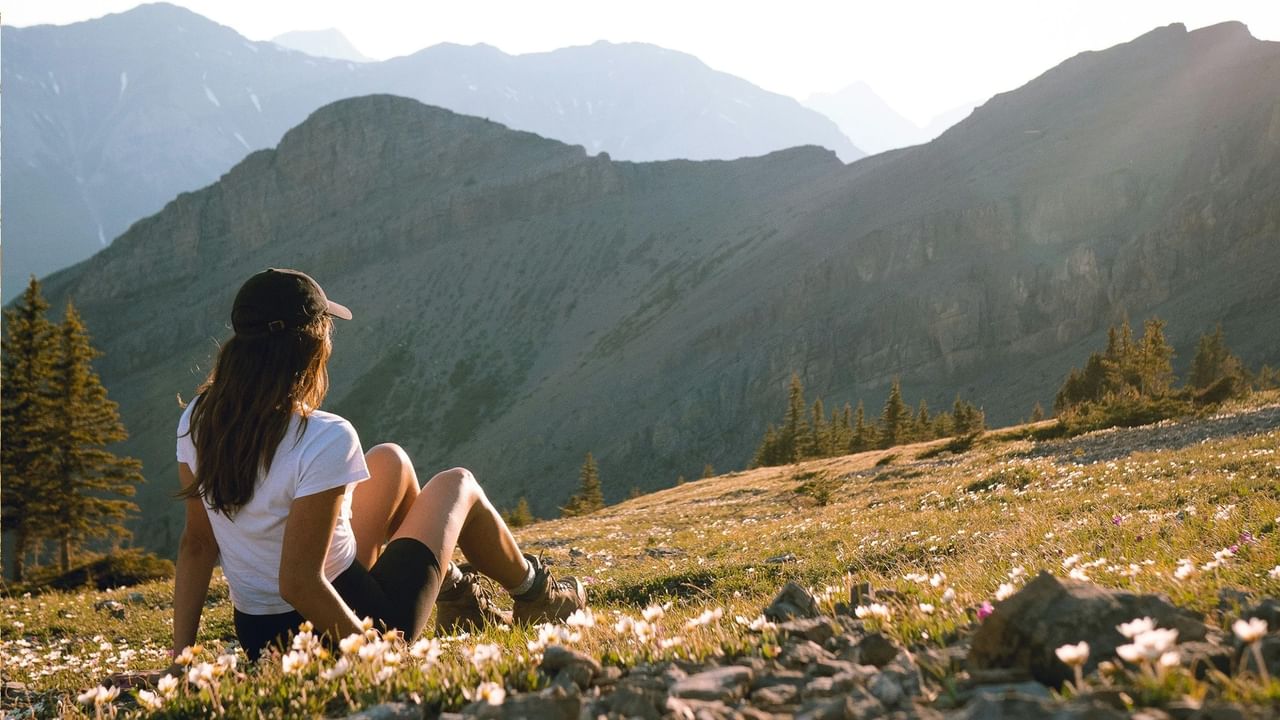Woman in white shirt and black hat sits on grassy mountain slope with wildflowers.