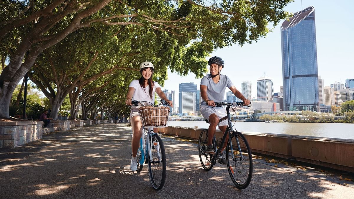 A couple is smiling while cycling along the scenic Brisbane River path with the city skyline near The Sebel Brisbane