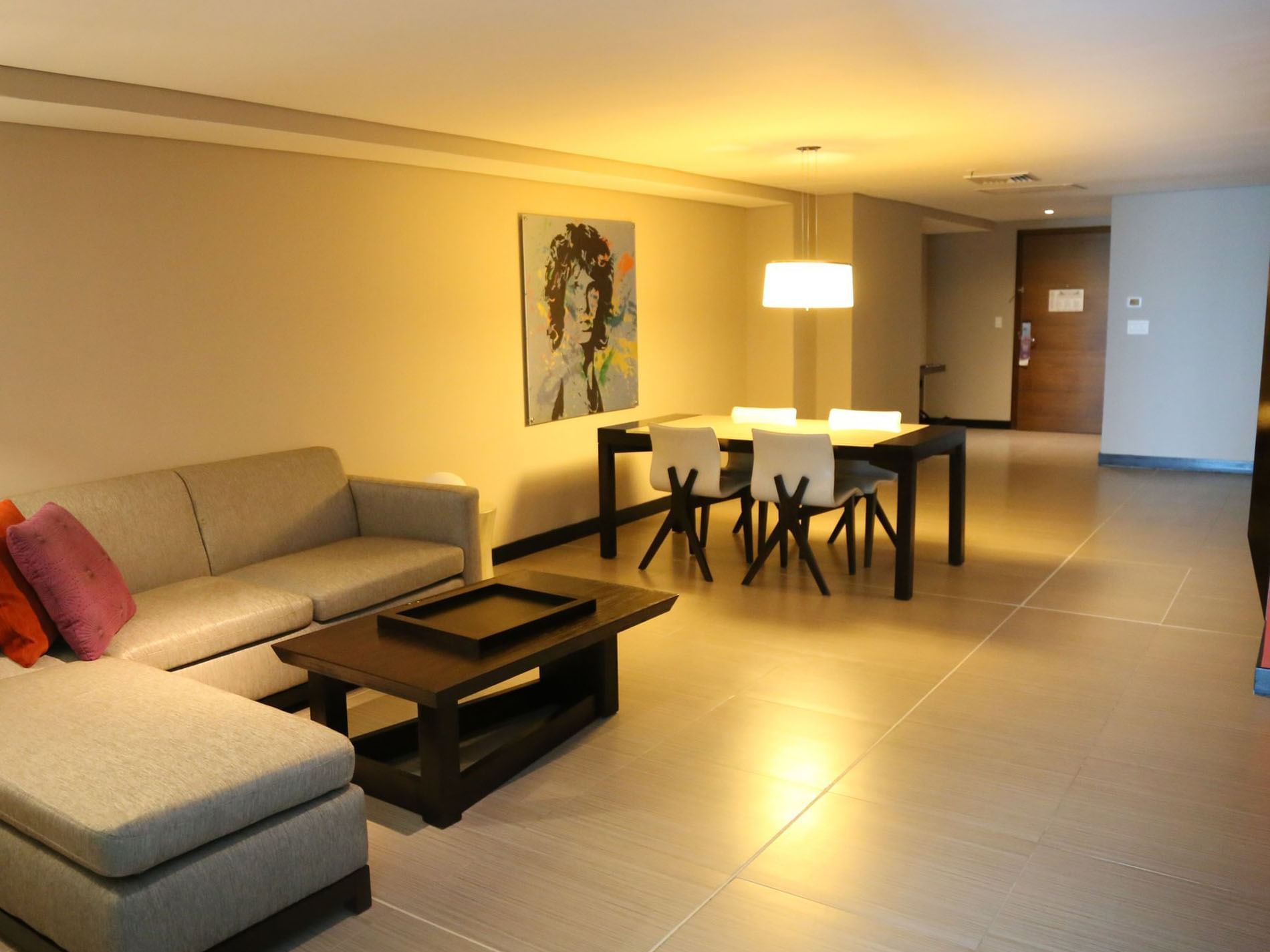 Hotel room with gray couch, coffee table, dining set, and contemporary painting on the wall.
