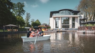 Family in boat on pond with modern building and gazebos on shore, trees around.