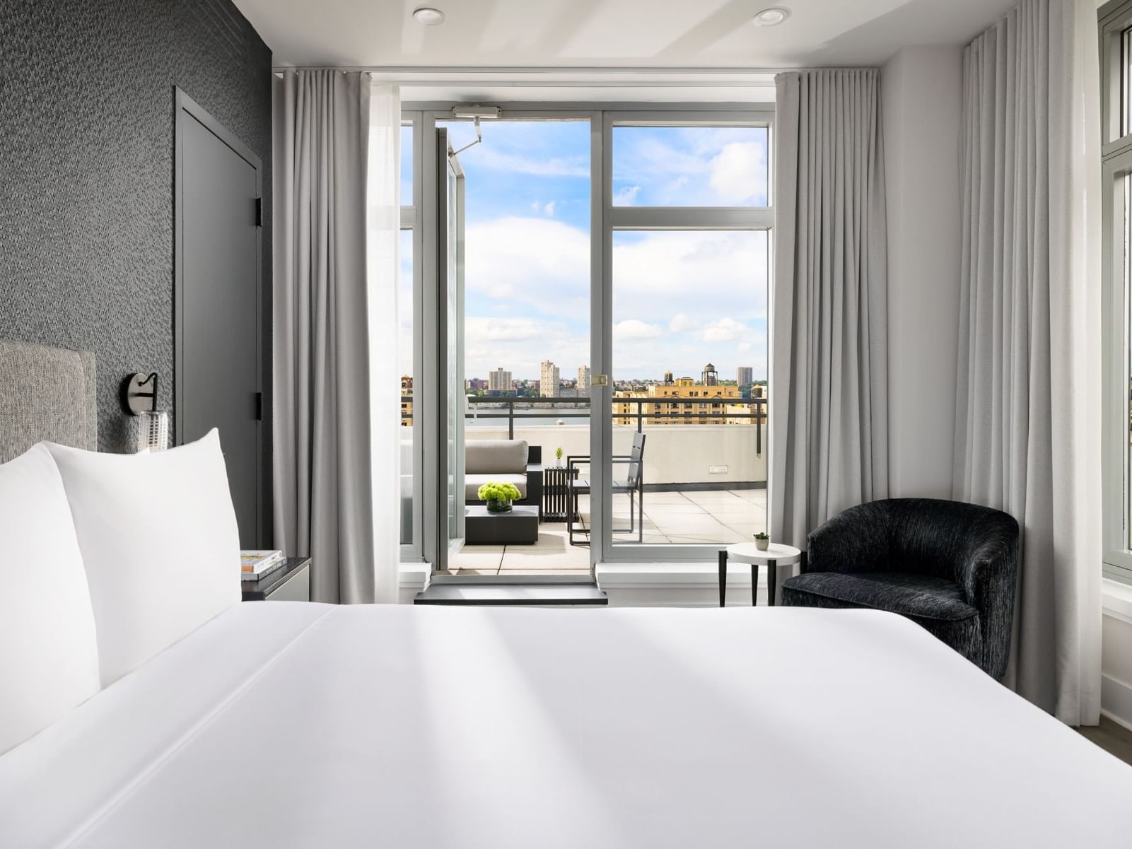 ArtHouse Hotel Skyline Penthouse with white bed, black chair, and open glass door leading to balcony with city view.