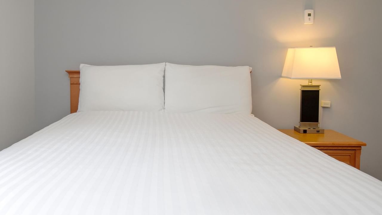 A made bed with two pillows and a lit lamp on a wooden nightstand beside it.