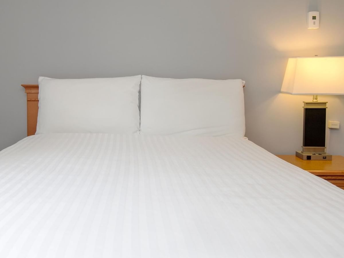 A neatly made bed with white bedding, two pillows, and a nightstand with a lit lamp.