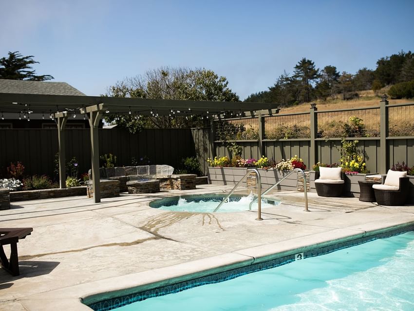 Outdoor pool and hot tub area with chairs, plants, and a fence under a clear blue sky.