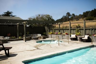 Outdoor pool and hot tub area with chairs, plants, and a fence under a clear blue sky.