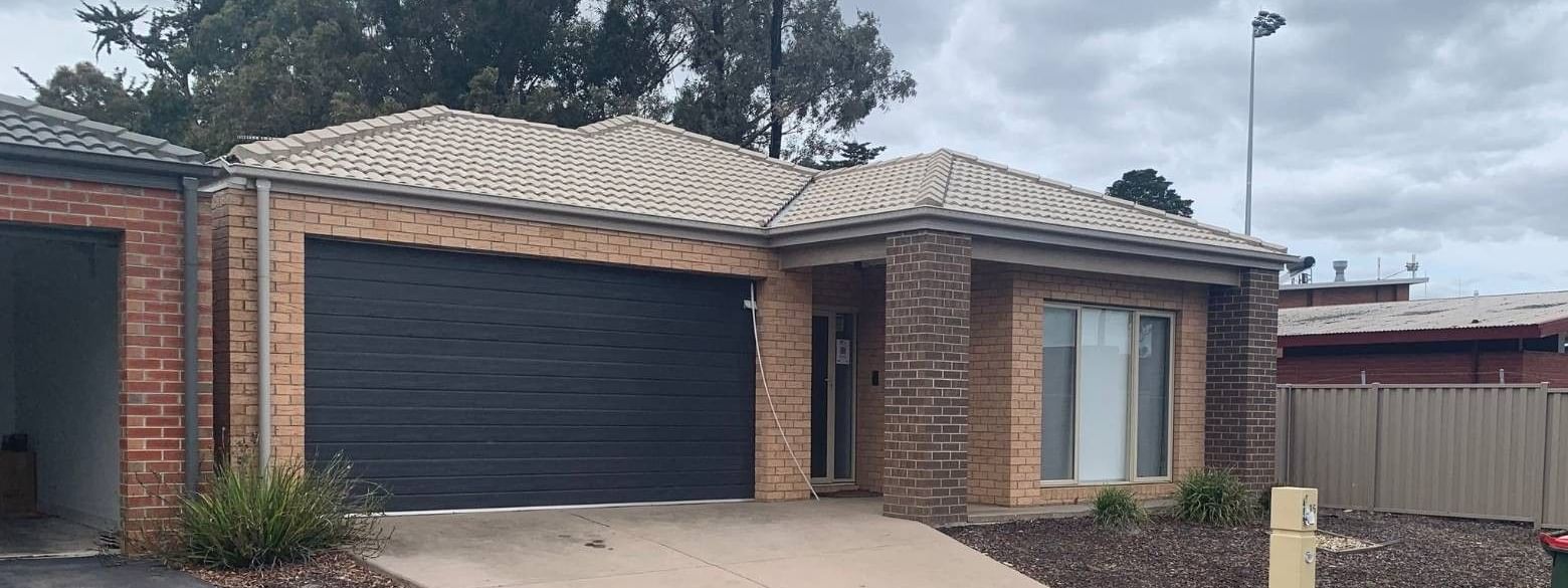 A brick house with a black garage door and driveway, surrounded by trees and fencing.