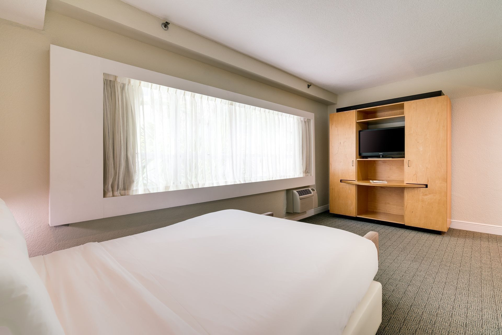 A wide view of a hotel room with a bed, window, TV cabinet, and carpeted floor.