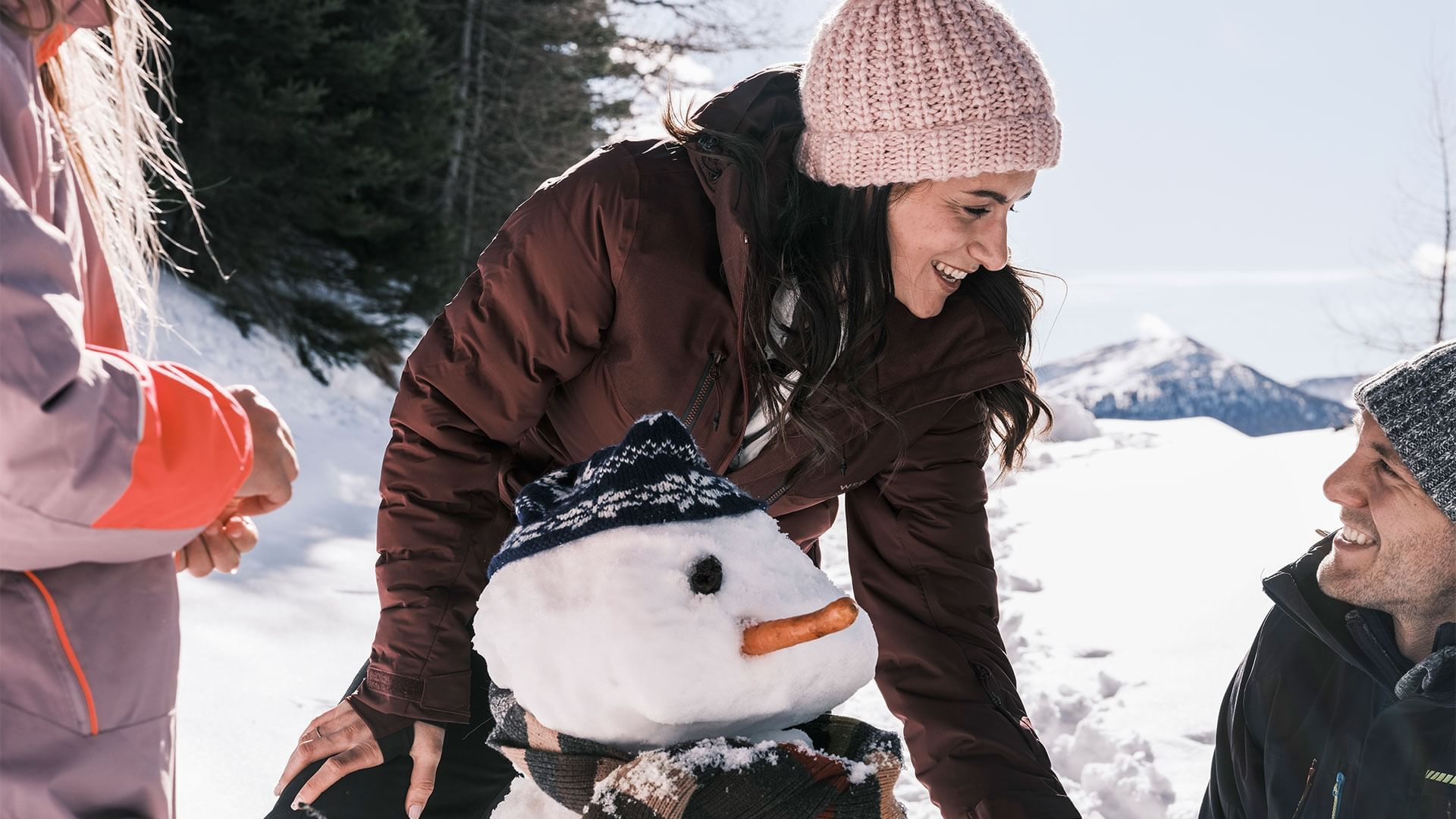 Eine Frau und ein Mann bauen im Schnee einen Schneemann, während ein Kind zuschaut.