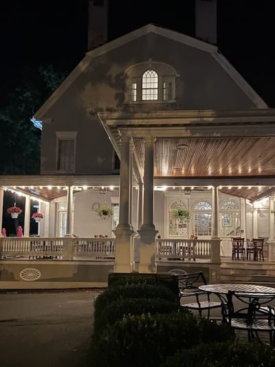 Outdoor porch with string lights and hanging flowers, by white columns near seating at night near Simsbury 1820 House