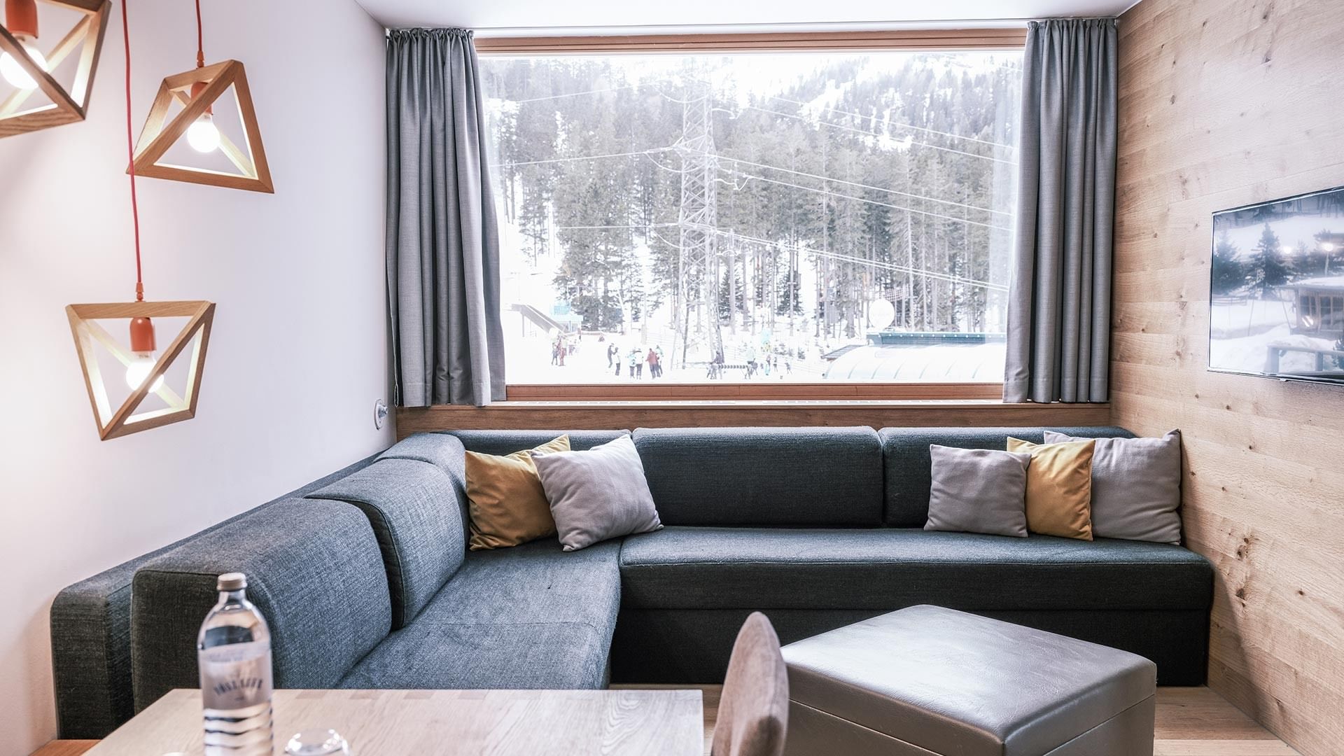 Wohnzimmer mit grauem Sofa, Kissen, Tisch, Fernseher und Fenster mit Blick auf verschneite Berge.