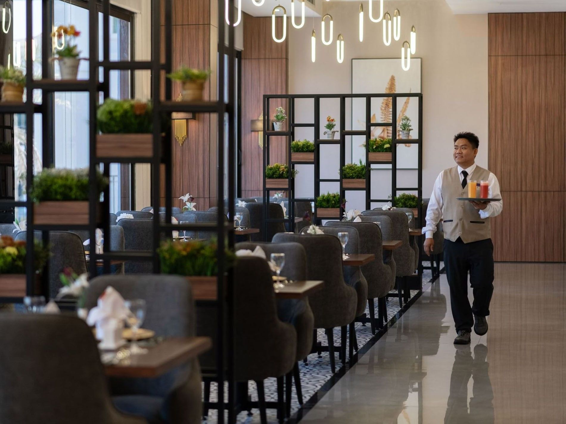Waiter carrying colorful juices through a dining area with gray chairs and metal shelving in La Maison at Warwick Al Jubail