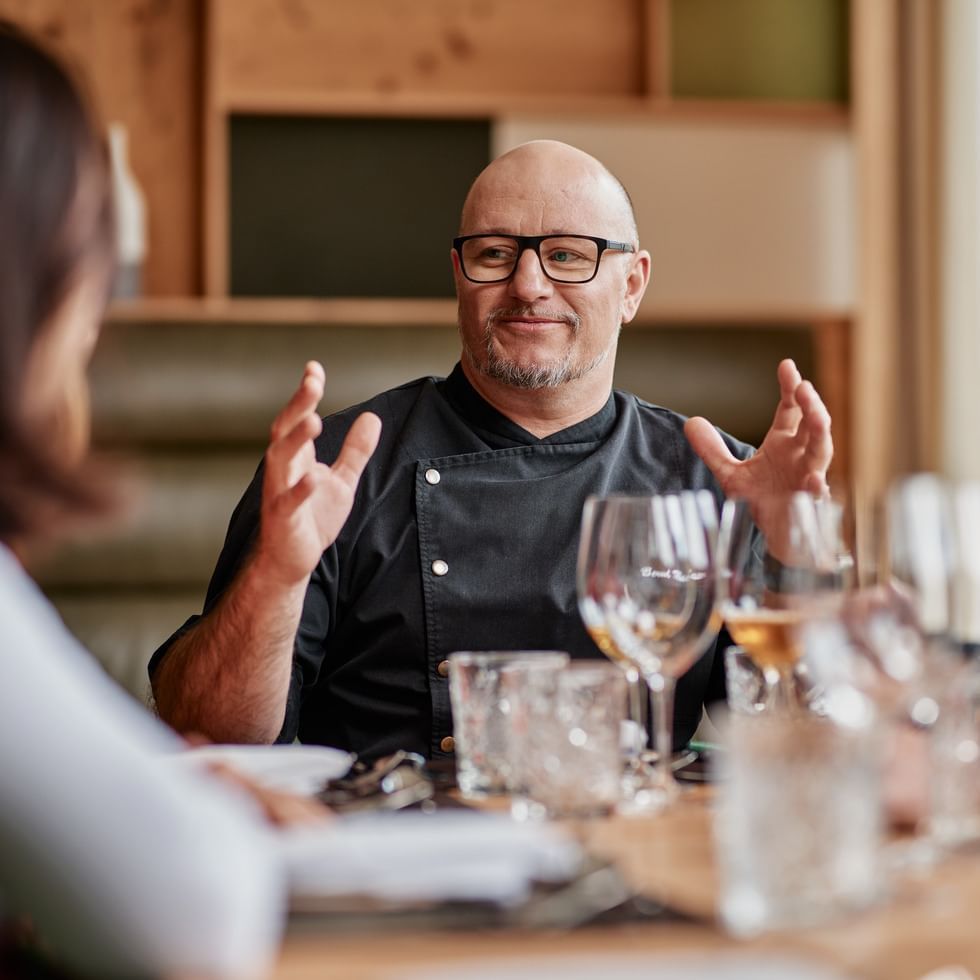 Uomo calvo in un ristorante, con occhiali e maglietta nera, gesticola davanti ad una donna.