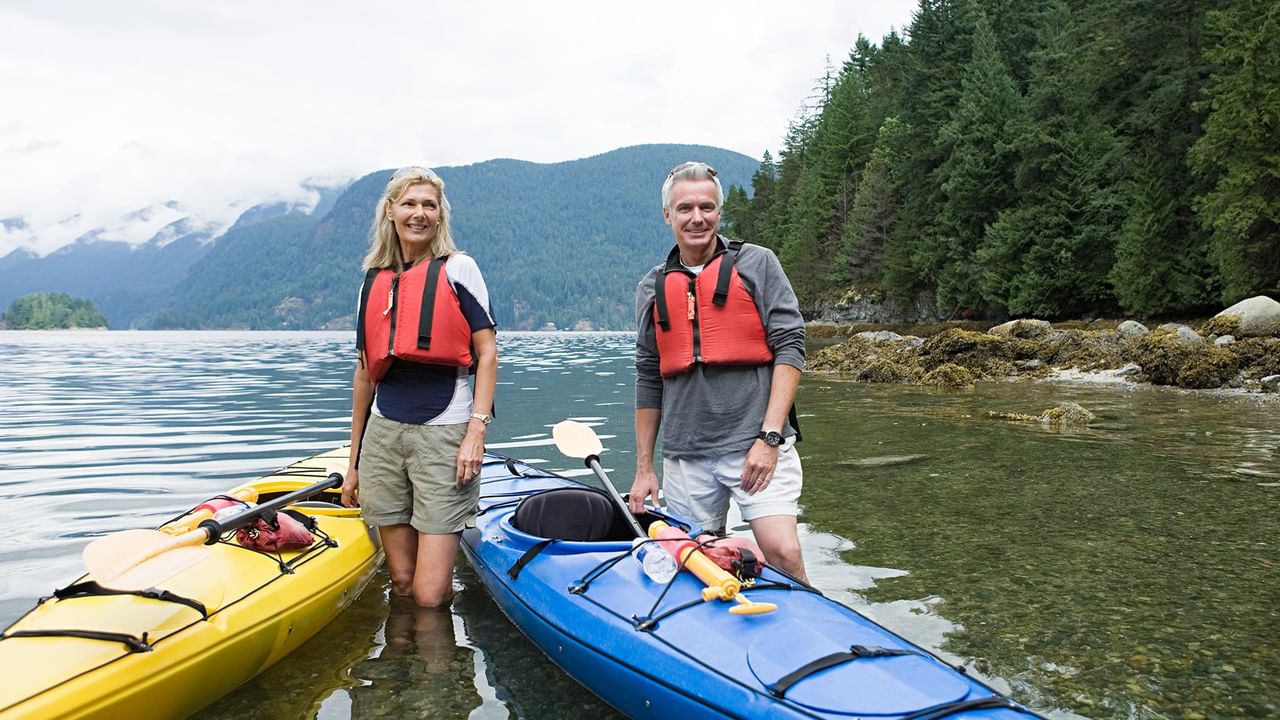 Two kayakers in red life jackets standing in shallow coastal water