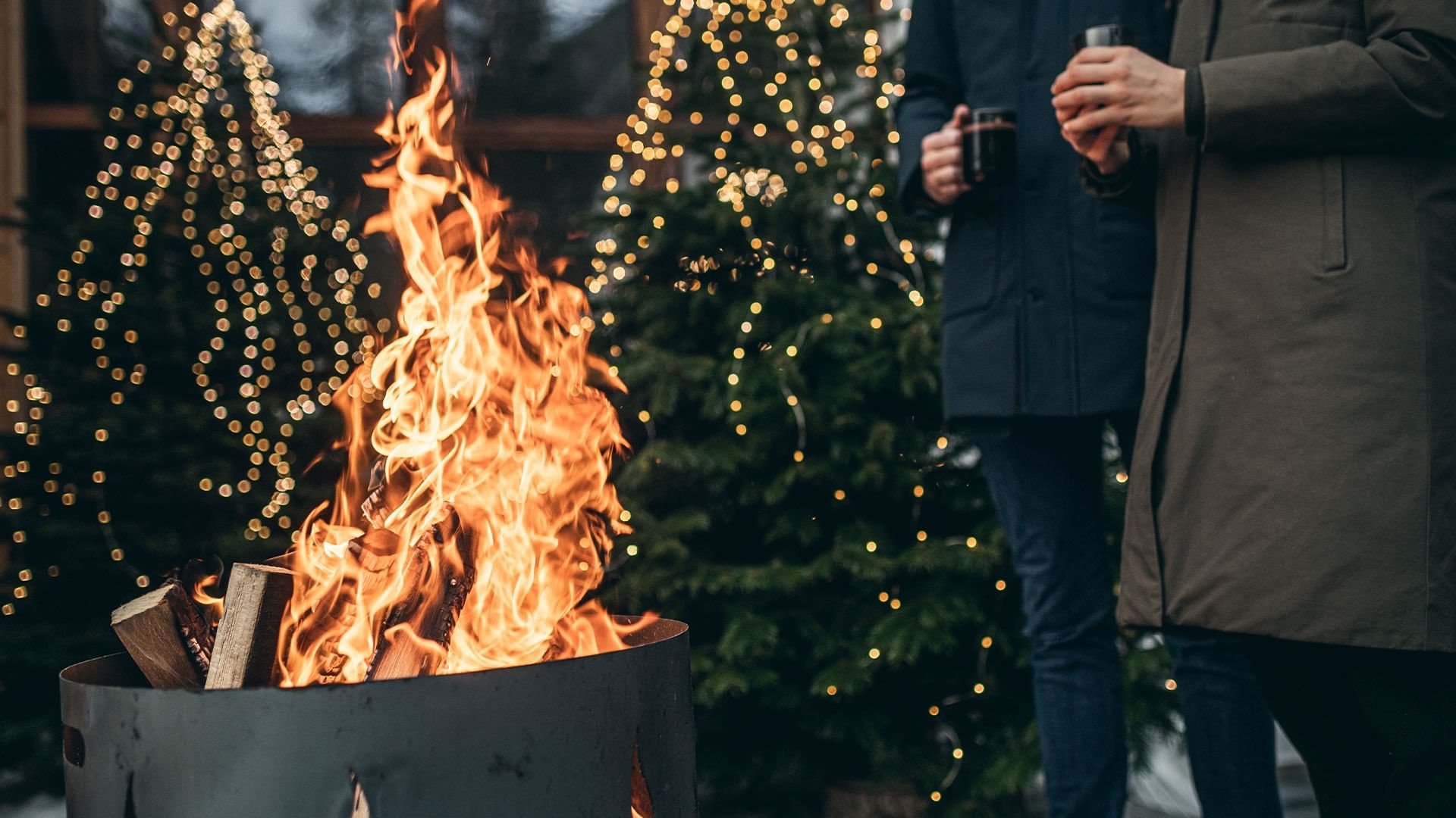 Zwei Personen mit Getränken vor Feuerschale und beleuchtetem Weihnachtsbaum.