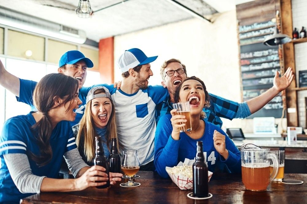 A group of friends in blue and white athletic gear hold beers and cheer at a bar.