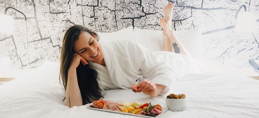 A person in a white hotel robe lounges on the bed and enjoys a charcuterie platter from the hotel.