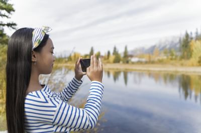 Girl taking a photo by a calm lake surrounded by autumn trees and distant mountains near Blackstone Mountain Lodge