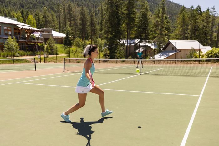Woman hitting tennis ball on court, with another player in background and mountain landscape.