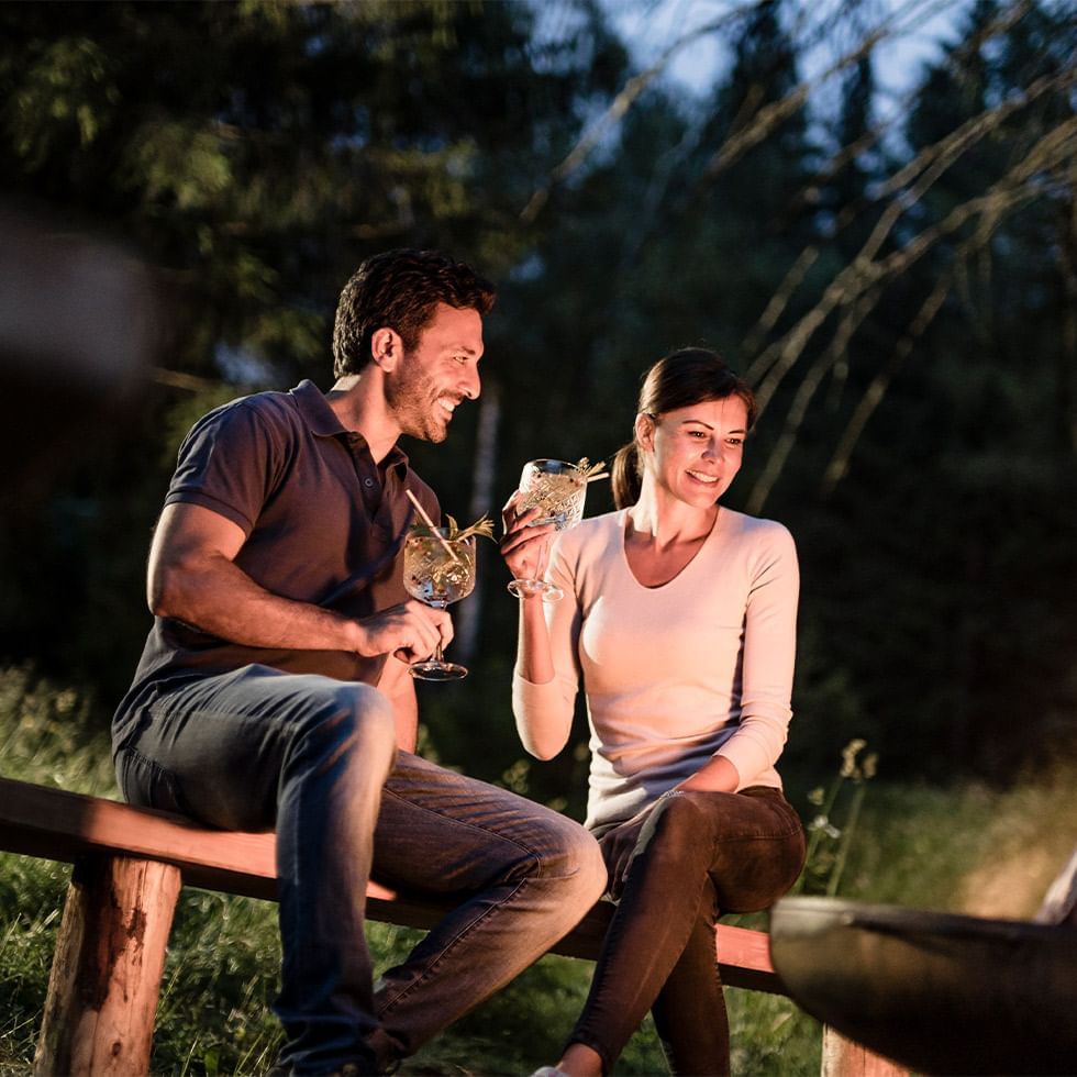 Couple on bench with drinks, smiling, near fire pit, forest backdrop at dusk.