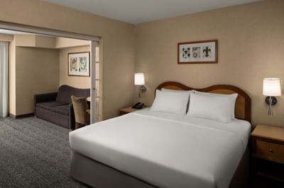 Bed by wooden nightstands, with a sofa through the doorway in the guest room at Anaheim Portofino Inn & Suites