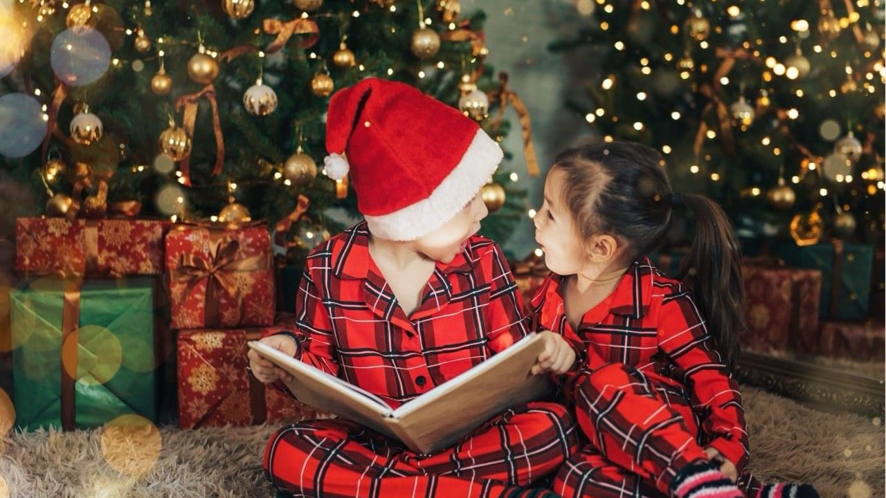 Two children in red pajamas sit by a Christmas tree, one reading a book, surrounded by gifts.