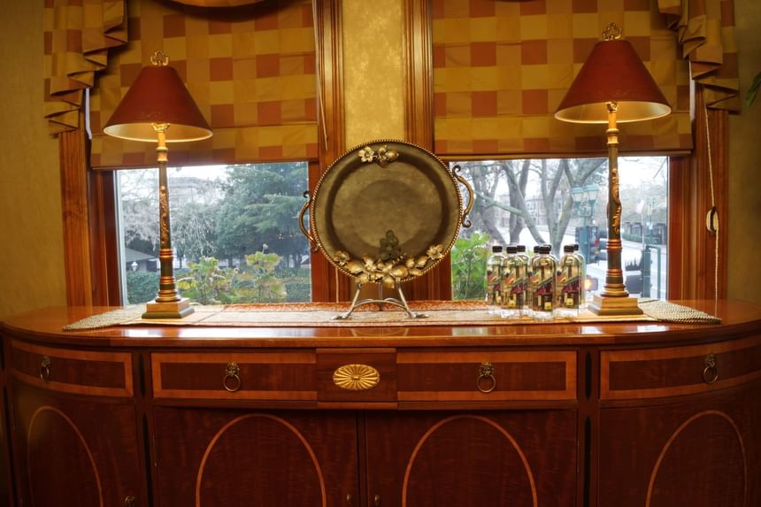 Wooden sideboard adorned with a decorative silver tray, lamps, and glass bottles, framed by large windows at The Rose Hotel