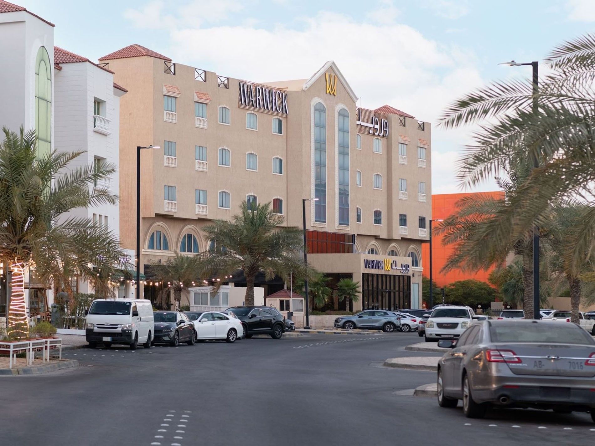 Multi-story exterior of Warwick Al Jubail by a paved street surrounding palm trees and a busy parking area
