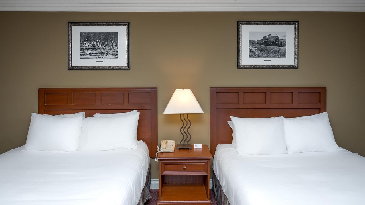 Two double beds with white linens, wooden headboards, shared nightstand with lamp, and framed artwork at Coast Edson Hotel.