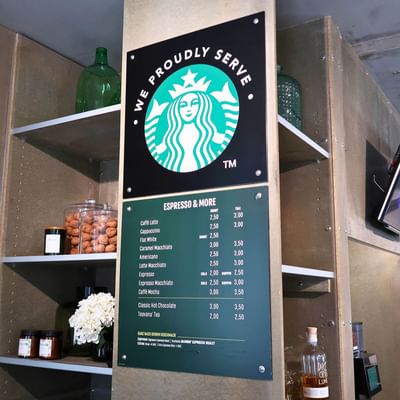 Starbucks menu board displaying espresso and beverage prices, alongside shelves with snacks at Avaneo Hotel