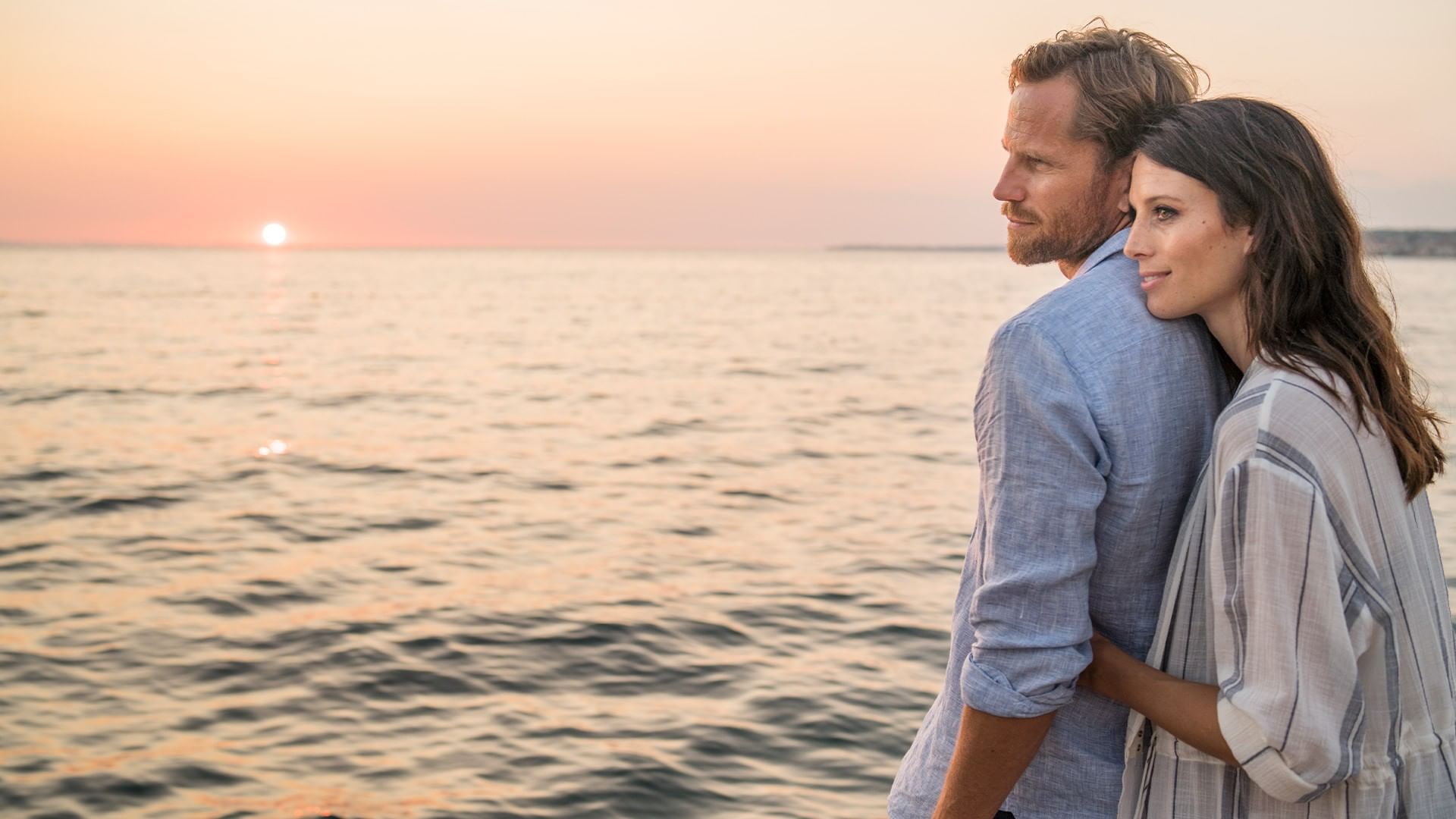 Couple standing by the sea at sunset, man with arm around woman's waist.