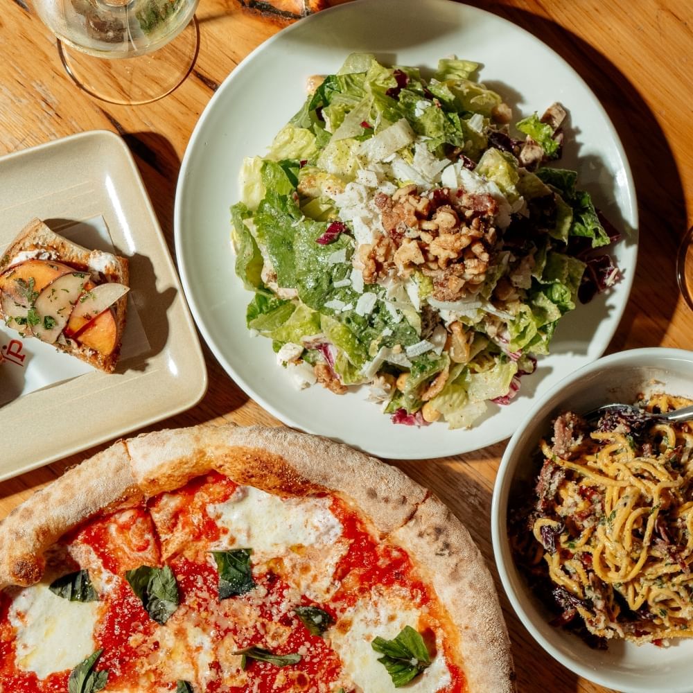 An  overhead view of a plate of salad, a pizza, a bowl of pasta, and a bruschetta on a wooden table. 