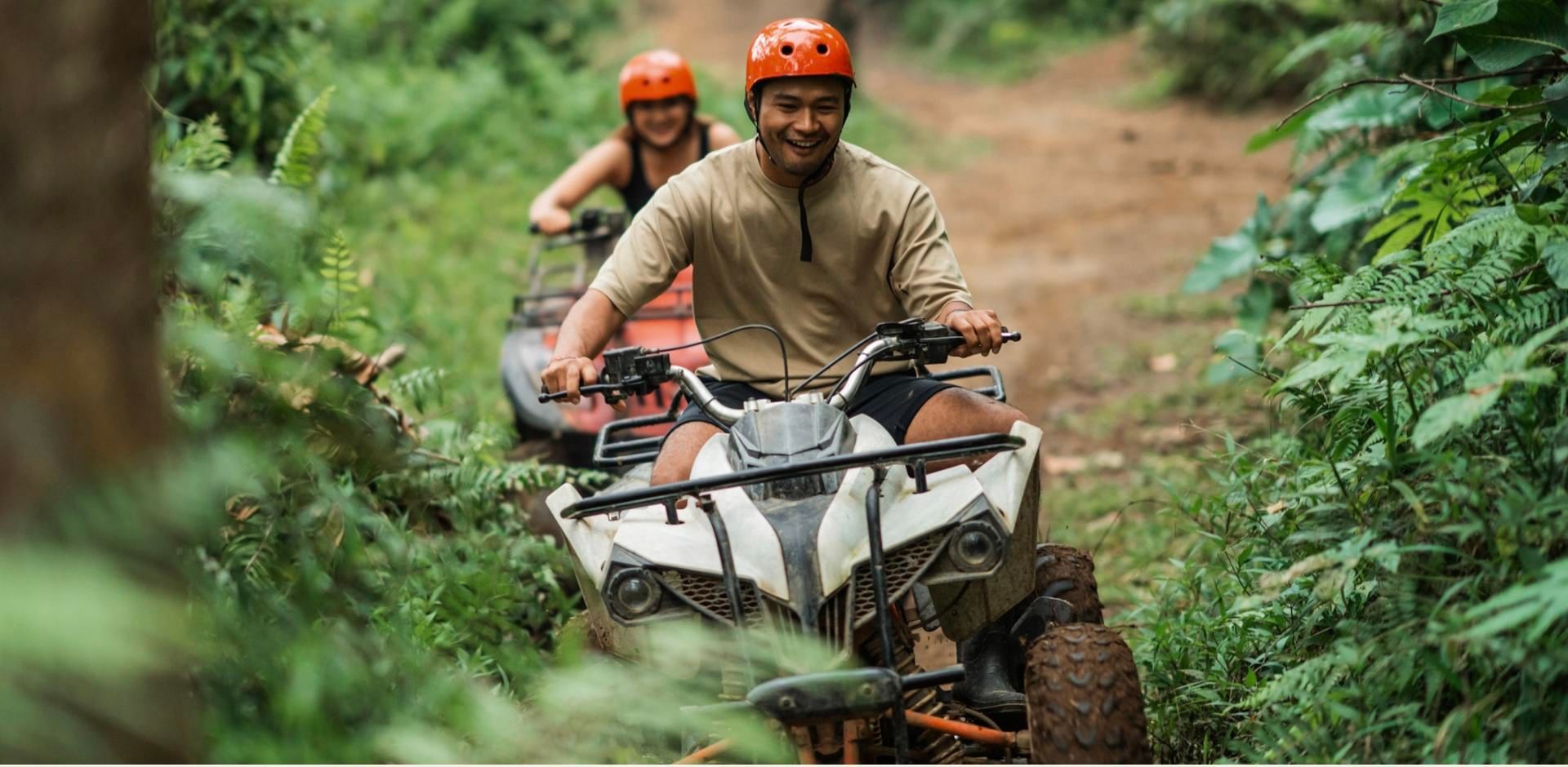 Two people in orange helmets ride white ATVs along a muddy path through lush green jungle near Tulum.