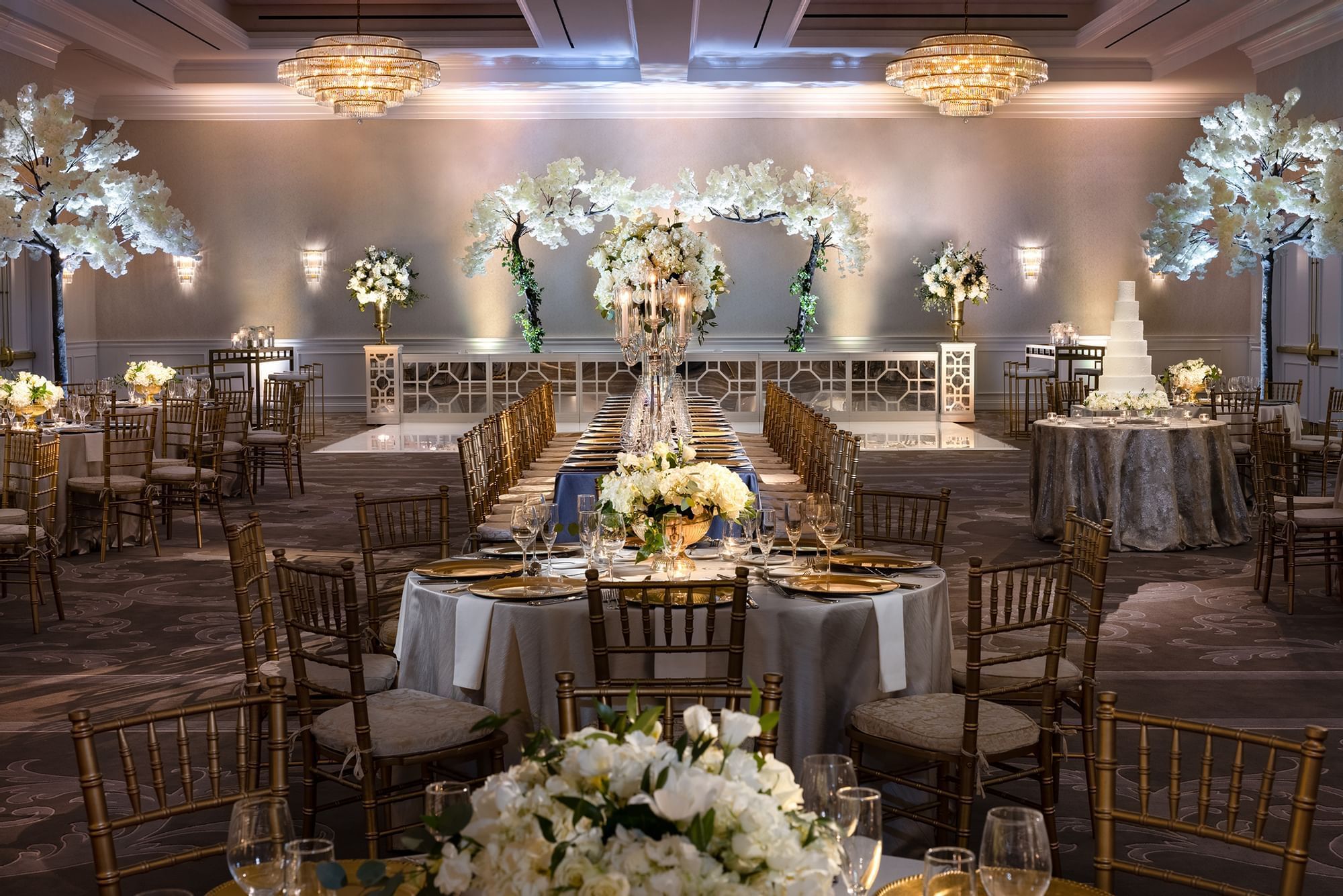 Long dining tables by floral trees under crystal chandeliers in ballroom at Warwick Melrose Dallas 