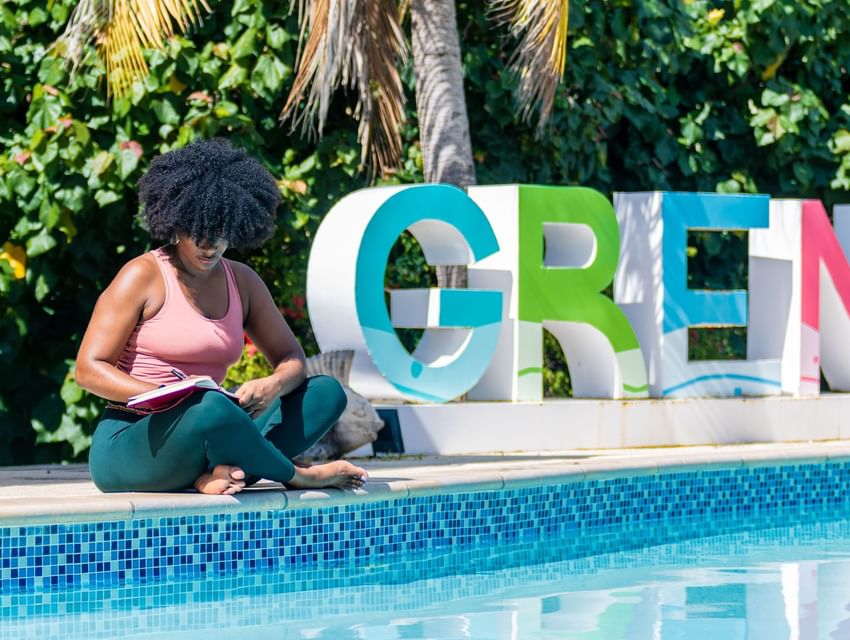 Woman writing by the pool with the Grenada sign in the background at True Blue Bay Hotel