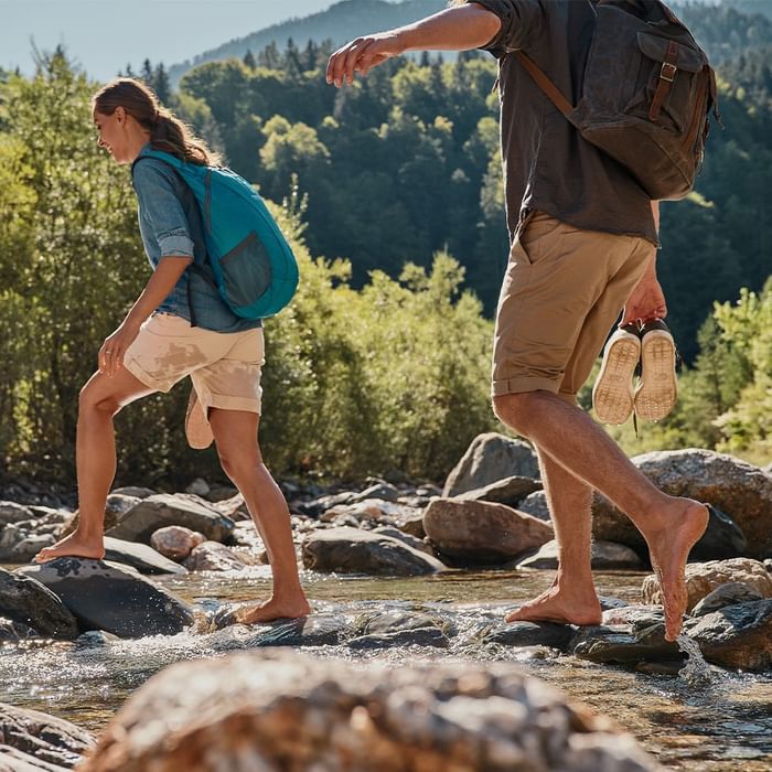 Two people crossing a stream by stepping on rocks, one holding shoes, both wearing backpacks.