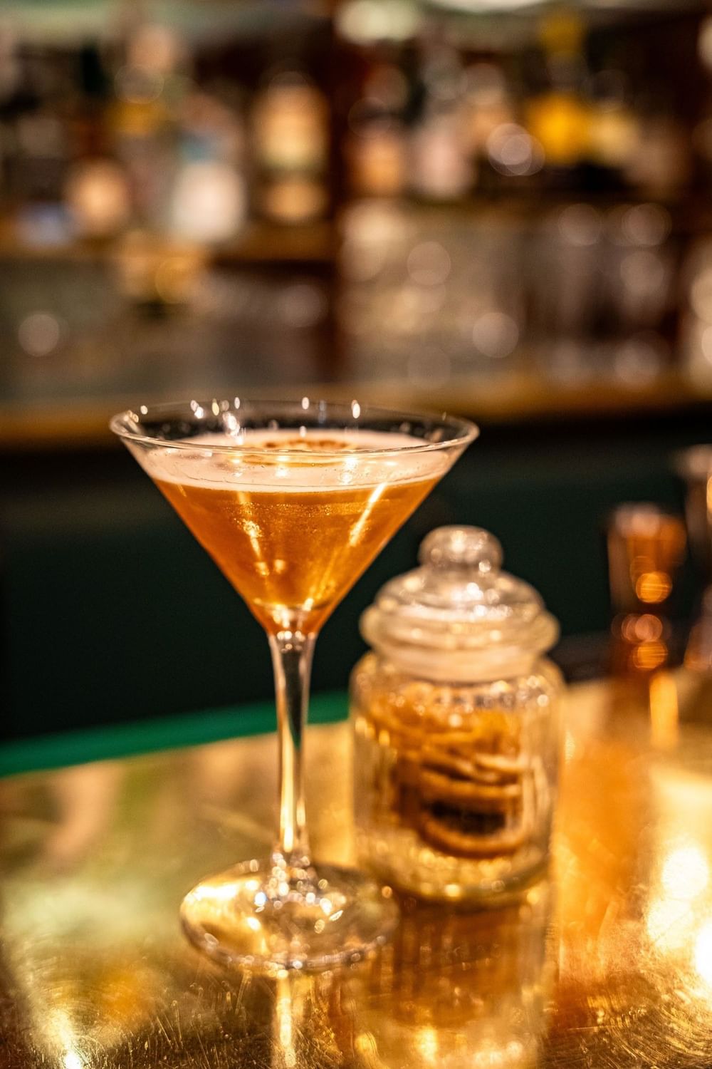Cocktail in a martini glass by a glass jar of garnishes on a golden counter served in Duke's Bar at Hotel Westminster Paris