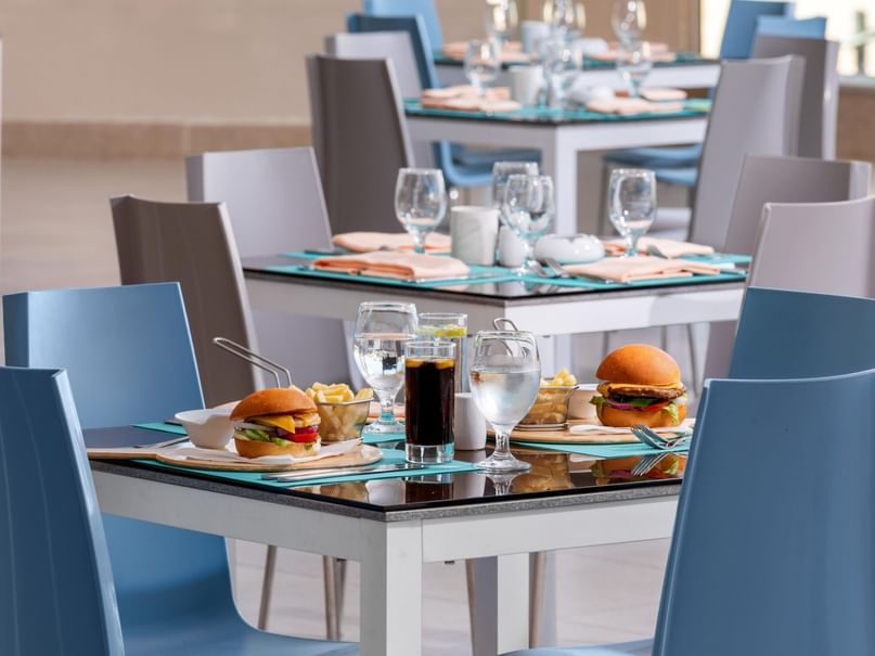 A restaurant with blue chairs, white tables, and two burgers with drinks and fries on the table.