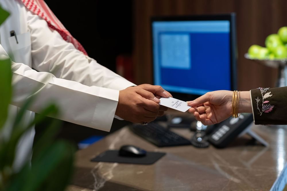 Receptionist handing a card to a guest by a computer monitor over a marble desk at Warwick Al Taif Hotel