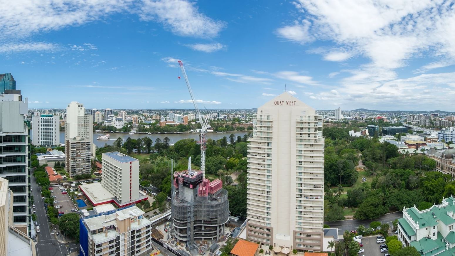 Aerial view of urban skyline with high-rise buildings, construction crane, river, and green parkland under blue sky.