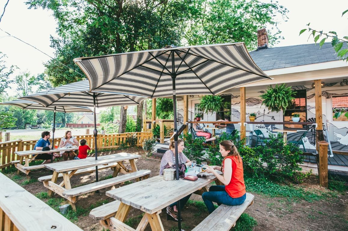 A group of people sitting at outdoor picnic tables under striped umbrellas.
