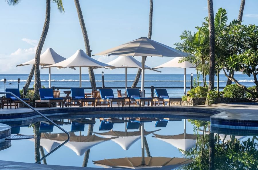 Blue lounge chairs by white umbrellas under palm trees surrounding a pool at Warwick Fiji Resort and Spa