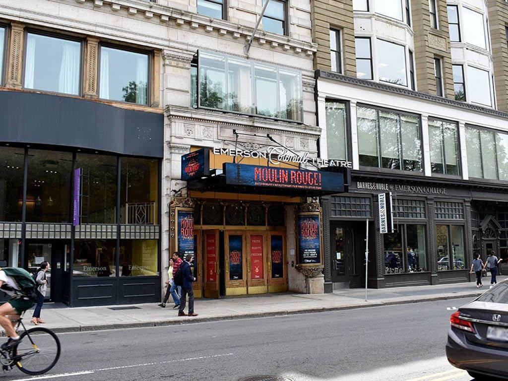 People walking on sidewalk in front of Emerson Colonial Theatre with Moulin Rouge! sign, shops, and passing vehicles.