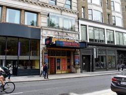 People walking on sidewalk in front of Emerson Colonial Theatre with Moulin Rouge! sign, shops, and passing vehicles.