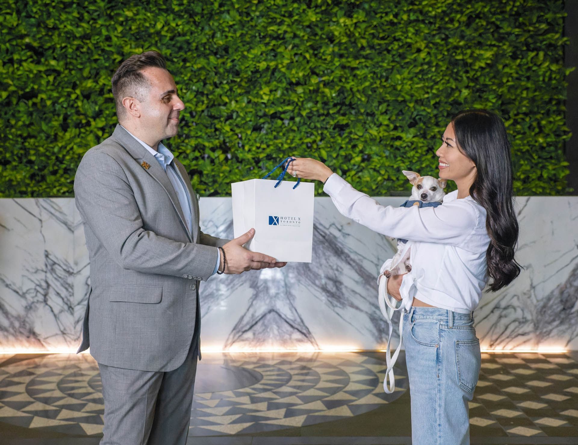 Staff member handing white gift bag to guest holding small dog beside marble wall and green plant at Hotel X Toronto