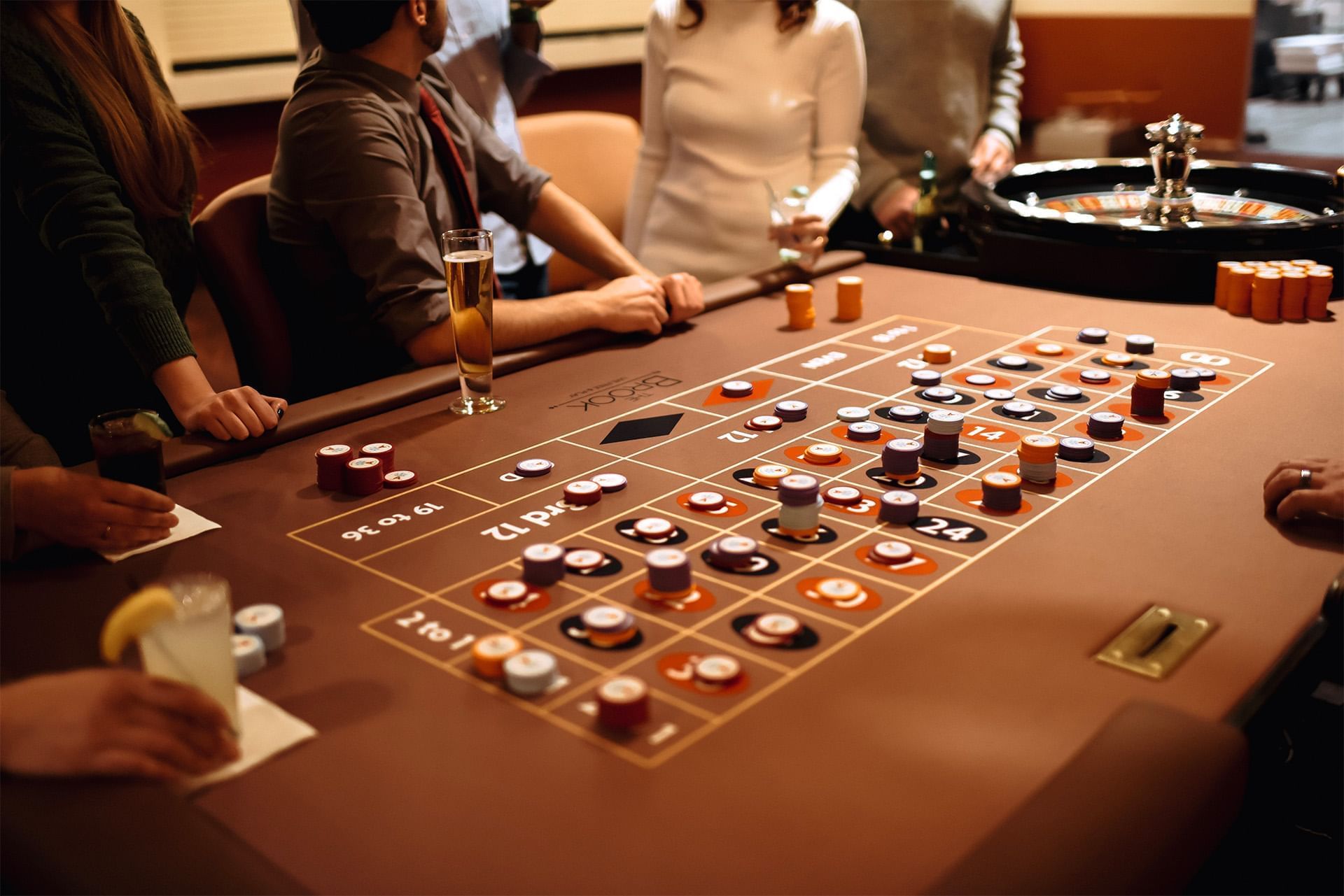 Guests gather by a casino roulette table with colorful betting chips and drinks under warm lighting at The Brook