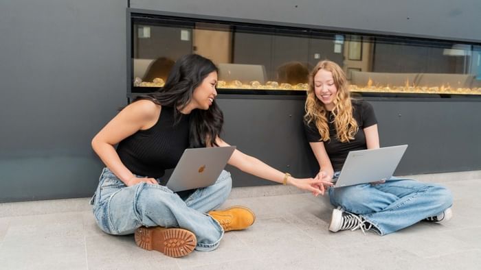 Two students are chatting and studying together on computers in front of a cozy fireplace. 