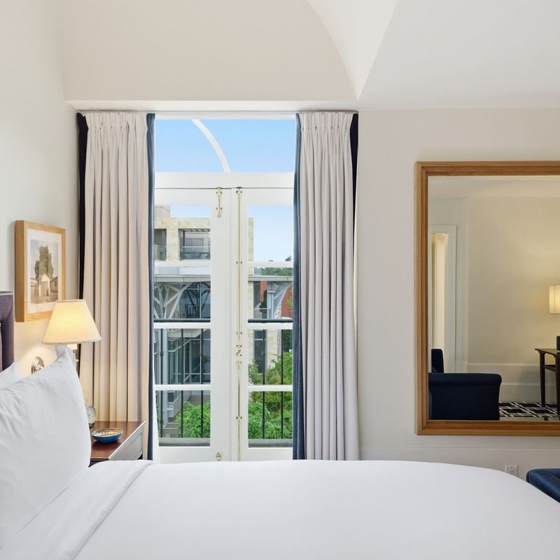 Bright hotel room with white bedding, tall windows with curtains, and a large mirror reflecting the adjacent space.