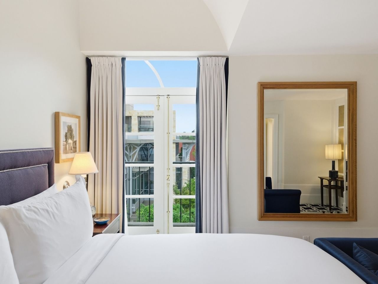 Bright hotel room with white bedding, tall windows with curtains, and a large mirror reflecting the adjacent space.