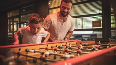 A man and a boy play a table soccer game in a room with a glass door.